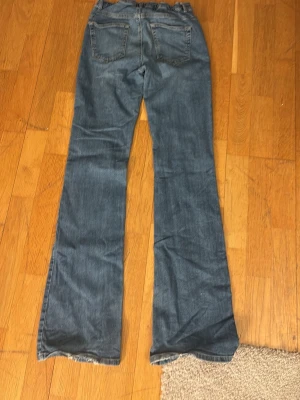 Blå bootcut jeans med slitna detaljer - Säljer ett par klassiska blå jeans med bootcut passform. Jeansen har en något utsvängd benvidd och femficksmodell. Materialet är robust denim i bomull och färgen är en tvättad blå ton. Perfekta för dig som gillar retrovibbar och avslappnad stil.