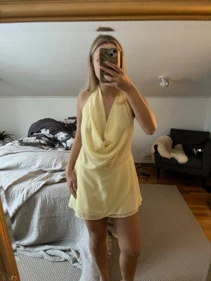 Ljusgul omlottklänning med öppen rygg - Superfin ljusgul kortklänning med omlott och draperad front. Klänningen har halterneck, öppen rygg med knyt och ett tunt, flowy material som känns luftigt. Perfekt för varma dagar och somriga tillfällen.