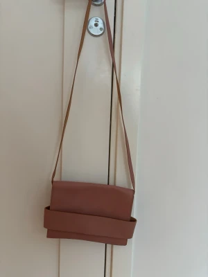 Dammrosa axelväska i skinn med magnetlås - Säljer en skitsnygg rosa axelväska i skinn med bred rem framtill. Väskan har ett magnetlås och insidan är beige. Perfekt storlek och enkel att matcha med olika outfits.