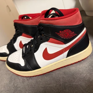 Nike Air Jordan 1 svart/röd/vit - Säljer ett par Nike Air Jordan 1 med klassisk svart, röd och vit färgkombination. Skorna har högt skaft, svart snörning och röd swoosh. Tillverkade i skinn med perforerad tåbox och retro-vibe. Perfekta för dig som gillar streetwear och sneakers med ikonisk design.