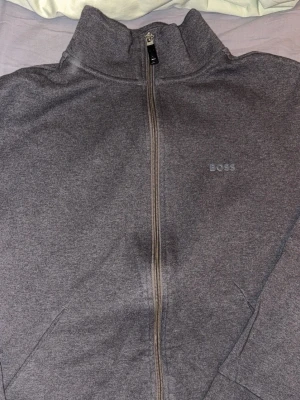 Grå ziptröja från BOSS - Säljer en stilren grå ziptröja från BOSS i storlek S. Tröjan har hög krage, hel dragkedja framtill och diskret BOSS-logga på bröstet. Perfekt till chill dagar eller när du vill ha en clean look. Materialet känns mjukt och bekvämt.