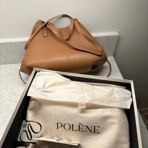 Polène Cyme väska i Camel - Säljer min jättefina väska från Polène i modellen Cyme. Färgen heter Camel. (”Stora”/vanliga storleken). Väskan har en avtagbar pouch som hänger på insidan av väskan! Måtten är 32 x 38 x 17.  Väskan har magnet-stängning i mitten och på sidorna och kan också stängas som på översta bilden i bild 5. Kan bäras i handen, i armvecket och över axeln. Ett par vattenfläckar finns på insidan av väskan. Miminala ”repor” som jag ej tycker syns på utsidan. Låda, etiketter och dustbag medföljer! 🤍🤍 