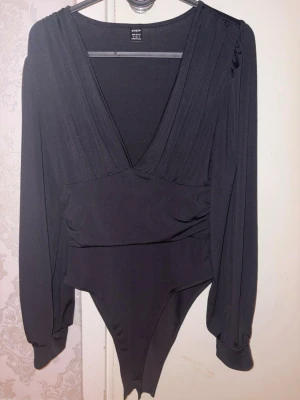 Svart omlott body från Shein - Säljer en svart body från Shein med djup v-ringning och omlottdetalj framtill. Långa ärmar med lätt puff och breda muddar. Stretchigt material som sitter snyggt på kroppen och ger en stilren look.