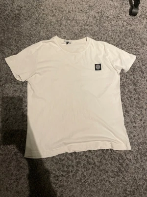 Vit t-shirt från Stone Island  - Denna märkes-T-shirt i vitt har en lätt urtvättad finish som ger ett avslappnat och trendigt uttryck. Tillverkad i mjuk och bekväm kvalitet som känns skön mot huden och håller passformen tvätt efter tvätt. Den diskreta vintagekänslan gör T-shirten enkel att matcha med både jeans och chinos, perfekt för en modern vardagsstil med en exklusiv touch.