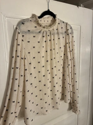 Vit/cremefärgad blus från Holly & Whyte - Vit/cremefärgad blus från Holly & Whyte by Lindex med små svarta rosettprickar. Lätt transparent, hög krage med volang och långa ärmar med smockade, volangkantade muddar. Romantisk och feminin stil.