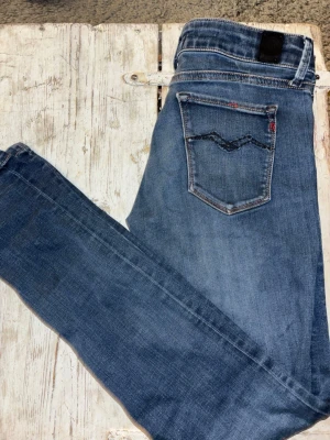 Blå Replay jeans - Säljer ett par klassiska blå jeans från Replay med raka ben och snygga slitningar. Jeansen har fem fickor, guldfärgade detaljer och tydliga sömmar. Materialet är mjukt jeans-tyg som sitter skönt och passar till det mesta. Perfekta för dig som gillar en tidlös look.