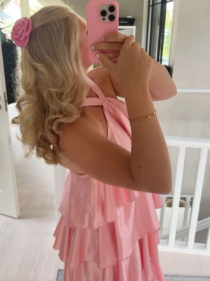 Rosa volangklänning med omlottband - Supersöt rosa långklänning med flera lager volanger och omlottband över axlarna. Klänningen har en mjuk glansig finish och är ärmlös, perfekt till balen. Materialet känns lätt och svalt mot huden.