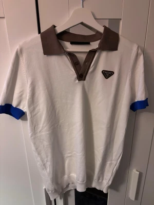 Vit polo Prada - Säljer en vit polotopp från Prada med brunt krage och knappslå samt blå detaljer vid ärmsluten. Klassisk och stilren design med Prada-logga på bröstet. 