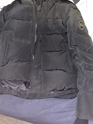 Svart pufferjacka från Canada Goose - Säljer en svart pufferjacka från Canada Goose med huva och diskret logga på ärmen. Jackan har dragkedja och tryckknappar framtill, samt ribbade muddar vid ärmsluten. Perfekt för kalla dagar och riktigt snygg till alla outfits.