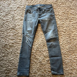 Dondup George jeans  - Säljer ett par grå dondup jeans med coola slitningar på bakfickorna. Jeansen har klassisk femficksdesign, låg midja och smal passform hela vägen ner. Perfekt för dig som gillar en urban och avslappnad stil.