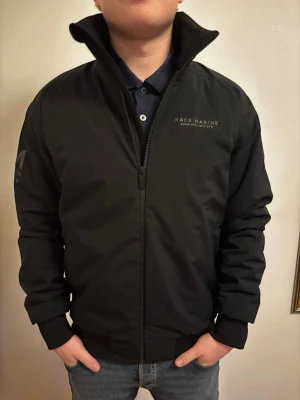 Svart vindjacka från Race Marine - Säljer en svart vindjacka från Race Marine med hög krage och dragkedja framtill. Jackan har diskreta loggor på bröstet och ena ärmen, samt två fickor med dragkedja. Perfekt för dig som vill ha en stilren och enkel jacka till vardags. Jackans ny pris är ungefär 1200 kronor köpt för 2 veckor sen men ej använt den. Var inte rädd för att pruta om mitt pris är för högt.