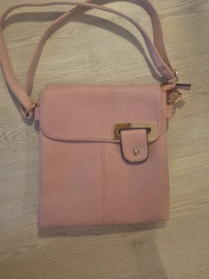 Rosa axelväska med gulddetaljer - Säljer en ljusrosa axelväska med justerbart axelband. Väskan har guldfärgade metalldetaljer, dragkedja på baksidan och ett lock med snygg knäppning framtill. Invändigt finns flera fack för bra organisering. Perfekt storlek för det viktigaste!