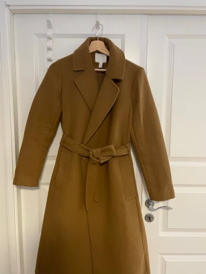 Kamelbrun kappa från H&M med bälte - Snygg kamelbrun kappa från H&M i klassisk modell med bred krage och knytskärp i midjan. Kappan är helfodrad och har två fickor framtill. Perfekt för höst och vinter när du vill vara både varm och stilren.