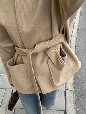 Beige omlottjacka med bälte - Säljer en beige omlottjacka med knytbälte i midjan och stora fickor framtill. Jackan har en rak, loose passform och är tillverkad i ett mjukt material som känns lyxigt. Perfekt för lager på lager och enkel att styla till jeans eller kjol.