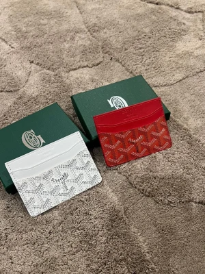  kortfodral i rött och vitt - Två stilrena kortfodral , ett i vitt och ett i rött. Båda har det klassiska mönstret och är tillverkade i exklusivt skinn. Perfekta för att hålla koll på korten med en lyxig touch och ikonisk design.