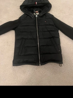 Svart pufferjacka Tommy Hilfiger XS - Säljer en svart pufferjacka från Tommy Hilfiger i storlek XS. Jackan har huva, dragkedja framtill och två fickor. Klassisk quiltad look med diskret logga på bröstet och detaljer i rött, vitt och blått på huvan. Perfekt för kyliga dagar.