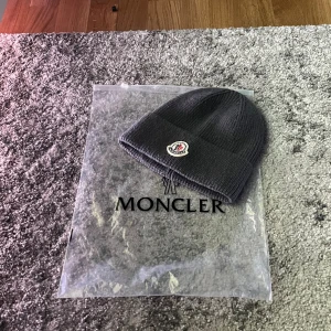Grå Moncler stickad mössa - Snygg grå stickad mössa från Moncler med klassisk logotyp framtill. Mössan har en ribbad design och uppvikt kant som ger en cool och enkel look. Perfekt för att hålla värmen under kyliga dagar och passar till de flesta outfits. Buda!