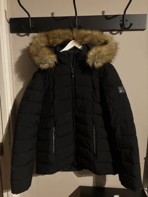 Svart pufferjacka med fuskpäls Mount Zero - Säljer en svart pufferjacka från Mount Zero i storlek L. Jackan har en mysig huva med fuskpäls, två fickor med dragkedja och quiltad design. Perfekt för kalla dagar och har en snygg, enkel look som funkar till det mesta.