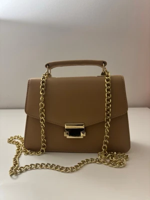 Beige handväska med guldkedja - Stilren beige handväska med guldfärgad kedja och metallspänne framtill. Väskan har ett topphandtag och en avtagbar axelrem i kedja, perfekt för att bära både i handen och över axeln. Klassisk och enkel design som lyfter vilken outfit som helst.
