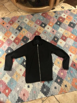 Svart overshirt med dragkedja - Säljer en svart overshirt från Weekday med hel dragkedja framtill och klassisk krage. Jackan är långärmad och har en clean, minimalistisk look som passar till det mesta. Materialet känns mjukt och lätt. Materialet är mocka! 