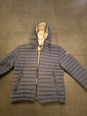 Blå dunjacka från Colmar - Säljer en helt oanvänd blå dunjacka från Colmar med huva och dragkedja framtill. Jackan har två sidofickor med dragkedja och quiltad design. Perfekt för kyliga dagar och riktigt snygg passform. Märke på ärmen och innerfoder i ljus färg. Säljes på grund av fel storlek