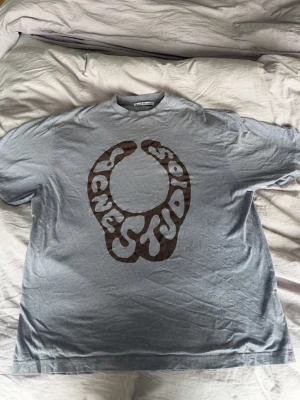 Grå Acne Studios t-shirt med tryck - Säljer en grå t-shirt från Acne Studios med ett stort brunt grafiskt tryck på bröstet där det står Acne Studios i en cool, vågig font. T-shirten har rund hals och korta ärmar. Perfekt för dig som gillar streetwear och vill sticka ut. Äkta och inga skambud