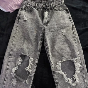 Grå slitna wide jeans med hål - Säljer ett par grå tvättade jeans med stora slitningar och hål på båda knäna. Byxorna har en loose fit med breda ben och klassisk femficksdesign. Råa kanter och distressed detaljer ger en riktigt edgy vibe. Perfekta för dig som gillar streetstyle.