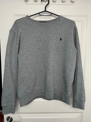 Grå sweatshirt från Polo Ralph Lauren - Klassisk grå sweatshirt från Polo Ralph Lauren med rund hals och broderad marinblå logga på bröstet. Tröjan har ribbade muddar vid ärmslut och nederkant, perfekt för en chill och stilren look. Tillverkad i mjuk bomullsmix för extra komfort.
