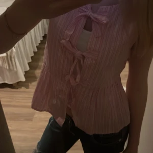 Rosa randig blus med rosetter Gina Tricot - Säljer en supersöt rosa blus från Gina Tricot i storlek XS. 💘Toppen är ärmlös, har ett randigt mönster i rosa och vitt samt tre dekorativa rosetter framtill. 🌸🎀.         Plagget är som en väst och passar då med ett linne/tröja under 😊