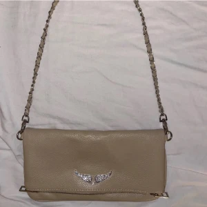 Beige axelväska med kedjerem - Säljer en beige axelväska i skinn med silverfärgad kedjerem och dragkedjedetaljer. OBS:äkta inte.Väskan har ett snyggt emblem med glittriga vingar på framsidan och en praktisk ficka med dragkedja på baksidan. Perfekt storlek för det viktigaste och ger en cool touch till din outfit.