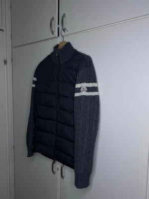 Grå Moncler stickad pufferjacka - Säljer en grå pufferjacka från Moncler med stickade ärmar och rygg i ull. Jackan har quiltad framsida i polyester, dragkedja och vita ränder på ärmarna samt Moncler-logga. Perfekt för dig som vill ha en unik och snygg jacka med sportig vibe. Akopia!!  Passar båda Moch L