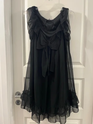 Svart omlottklänning med volanger - Säljer en svart omlottklänning med volangdetaljer och stor rosett framtill. Klänningen är ärmlös och har ett lätt, transparent tyg med volanger längs kanterna. Perfekt för dig som vill ha en luftig och söt look.