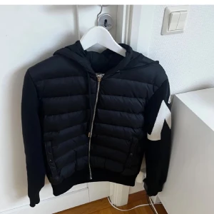 Svart Moncler pufferjacka med huva - Säljer en svart pufferjacka från Moncler med huva och dragkedja framtill. Jackan har ett stort vitt zigzag-mönster på ärmen och Moncler-logga. Fylld med dun och har ribbade muddar. Perfekt för dig som vill ha en snygg och varm jacka med streetkänsla.