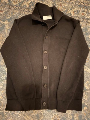 Svart kofta från Selected Homme - Stilren svart kofta från Selected Homme med klassisk krage och knäppning framtill. Tröjan är långärmad och har ribbade muddar vid ärmslut och nederkant. Perfekt för lager på lager och enkel att matcha till olika outfits.