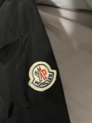 Svart Moncler dunjacka - Säljer en svart Moncler dunjacka med klassisk logga på ärmen och coolt serietidningsfoder på insidan. Jackan har dragkedja och är vadderad för extra värme. Perfekt för dig som vill ha en stilren och exklusiv vinterjacka.