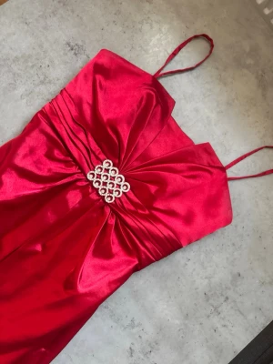 Röd långklänning med strassdetalj - Snygg röd långklänning i glansigt material med tunna axelband och en stor dekorativ rosett framtill, prydd med strass. Klänningen har en elegant v-ringning och markerad midja som ger en riktigt festlig look.