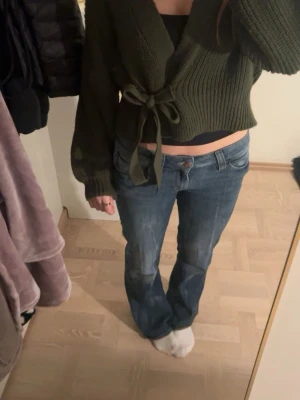 Mörkgrön stickad omlottkofta - Säljer en mörkgrön stickad omlottkofta med knytband i midjan. Koftan har breda, långa ärmar och ribbade muddar. Croppad modell som ger en snygg siluett och passar perfekt till jeans eller kjol.