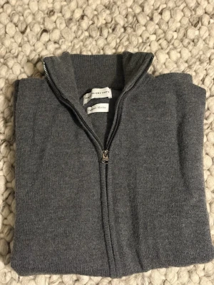 Grå merinoull kofta från The Glory Days - Mörkgrå kofta i 100% merinoull från These Glory Days. Klassisk modell med hel dragkedja framtill och hög krage. Perfekt för lager-på-lager och riktigt skön att ha på sig under kyliga dagar. Stilren och enkel design som funkar till det mesta.