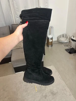 Svarta höga mockastövlar med dragkedja - Säljer ett par svarta, höga stövlar i mjuk mocka med grov sula och rund tå. Stövlarna har dragkedja baktill med snygga detaljer i metall och läder. Perfekta till höst och vinter för en stilren look.Äkta mocka, fodrad med päls.