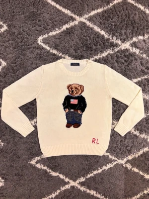 Polo Bear Ralph Lauren Vit - Säljer en ikonisk vit stickad tröja från Polo Ralph Lauren med den klassiska Polo Bear på bröstet. Tröjan har rund hals, långa ärmar och RL-broderi i rött nedtill. Jag väger 70KG och är 180CM och den passar mig inte alls 