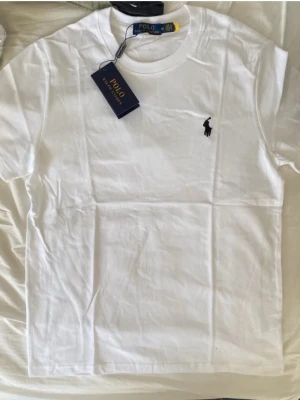 Vit t-shirt från Polo Ralph Lauren - Säljer en fräsch vit t-shirt från Polo Ralph Lauren med klassisk rund hals och korta ärmar. T-shirten har den ikoniska svarta Polo-loggan broderad på bröstet och är gjord i mjuk bomull. Perfekt för en clean och stilren look.