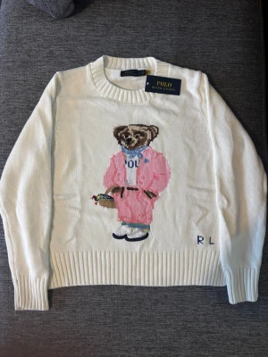 Vit stickad tröja Polo Ralph Lauren - Vit stickad tröja från Polo Ralph Lauren med ikonisk björn i rosa outfit broderad på framsidan. Tröjan har rund hals, ribbade muddar och är långärmad. Tillverkad i 100% bomull och har en relaxed vibe med snygga detaljer.