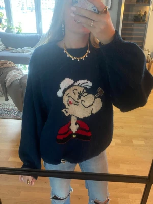 Mörkblå stickad tröja med Popeye - Säljer en mörkblå stickad tröja med stor Popeye-tryck på framsidan. Tröjan har en loose passform och långa ärmar, perfekt för dig som gillar coola och unika motiv. Materialet är mjukt och skönt, passar dig som vill ha något extra till hösten eller vintern.