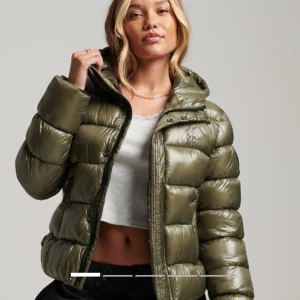 Olivgrön pufferjacka med dragkedja - Säljer en olivgrön pufferjacka med glansig finish och hög krage. Jackan har en klassisk quiltad design och stängs med dragkedja framtill. Perfekt för kyliga dagar och ger en cool streetstyle-vibe.