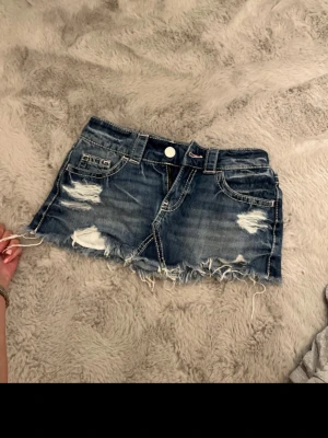 Blå jeanskjol med slitningar - Säljer en kort jeanskjol i blå denim med slitna detaljer och fransig nederkant. Kjolen har klassiska fickor fram och bak, bälteshällor och knappstängning. Perfekt för dig som gillar en edgy och avslappnad stil.
