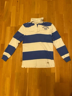 Randig rugbytröja Peak Performance - Säljer en klassisk rugbytröja från Peak Performance med breda blå och vita ränder, broderad logga på bröstet och stort '86' på ryggen. Tröjan har lång ärm, krage och är gjord i mjuk bomull. Perfekt för en sportig och avslappnad stil.