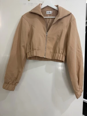 Beige croppad bomberjacka - Säljer en beige croppad bomberjacka från Pretty Little Thing. Jackan har dragkedja framtill, bred krage och resår i midjan samt vid ärmsluten. Perfekt för dig som gillar en avslappnad men trendig look.