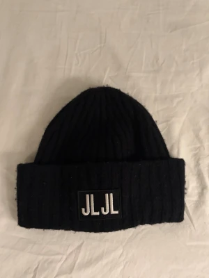 Mössa från J.Lindeberg - Säljer en svart ribbad mössa från J.Lindeberg med JLJL-logga framtill. Mössan är tillverkad i ull och är i bra skick. 
