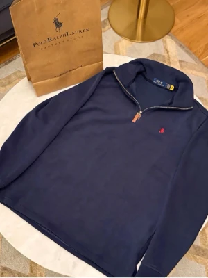 Marinblå half zip från Polo Ralph Lauren - Säljer en marinblå långärmad tröja från Polo Ralph Lauren med klassisk röd logga broderad på bröstet. Tröjan har en snygg half zip-dragkedja med läderdetalj och är gjord i mjuk bomull. Perfekt för dig som gillar stilrena och tidlösa plagg.