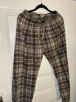 Rutiga pyjamasbyxor Rise and Shine - Rutiga pyjamasbyxor från Rise and Shine Nightwear collection. Byxorna har dragsko i midjan och är i mjukt bomullsmaterial. Färgerna går i brunt, beige och grönt med klassiskt rutmönster. Perfekta för chill dagar hemma eller som pyjamasbyxor. Jag kommer att sälja en tillhörande nattskjorta separat.